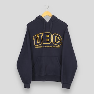 May include: Navy blue hooded sweatshirt with a kangaroo pocket. The front features the yellow text "UBC" above "UNIVERSITY OF BRITISH COLUMBIA". The sweatshirt is hanging from a wooden hanger.