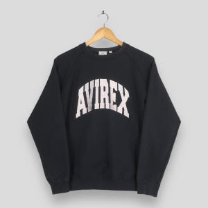 May include: A black long-sleeve sweatshirt featuring the word AVIREX printed in a large, arched white font across the chest. The garment has a crew neckline, raglan sleeves, and ribbed cuffs and hem, displayed on a wooden hanger.
