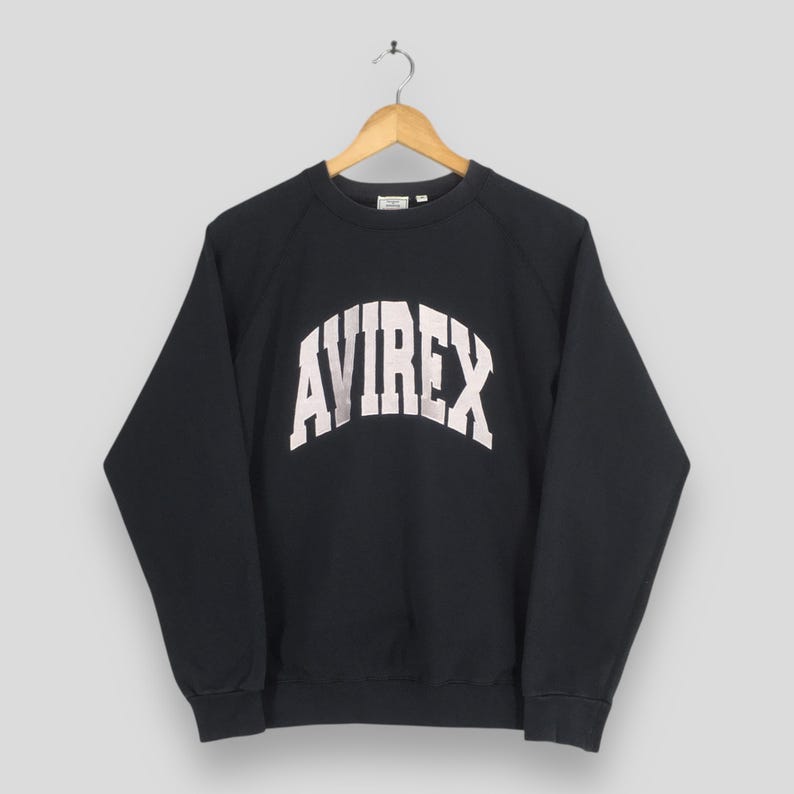 May include: A black long-sleeve sweatshirt featuring the word AVIREX printed in a large, arched white font across the chest. The garment has a crew neckline, raglan sleeves, and ribbed cuffs and hem, displayed on a wooden hanger.