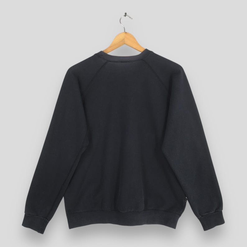 May include: A solid black long-sleeve sweatshirt featuring a crew neckline and raglan sleeves. The garment is displayed on a wooden hanger against a plain light gray background, showcasing a simple design with ribbed cuffs and a ribbed hemline.