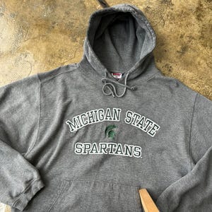Puede incluir: Sudadera con capucha gris con las palabras "MICHIGAN STATE SPARTANS" en letras blancas y verdes. Tiene un bolsillo delantero y una capucha con cordón. El logotipo de Spartan es verde.