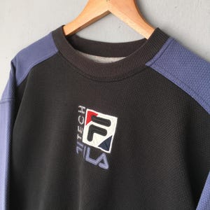 May include: Black and blue FILA Tech sweatshirt with a textured pattern. The sweatshirt has a crew neck and features the FILA logo in white, red, and blue. The sleeves are blue, and the sweatshirt is hanging on a wooden hanger.
