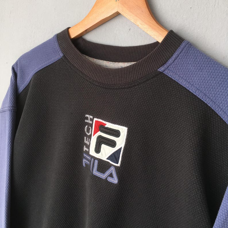 May include: Black and blue FILA Tech sweatshirt with a textured pattern. The sweatshirt has a crew neck and features the FILA logo in white, red, and blue. The sleeves are blue, and the sweatshirt is hanging on a wooden hanger.