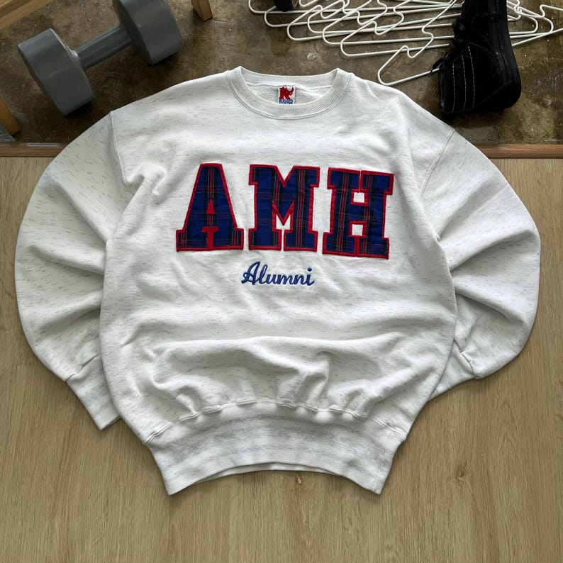 May include: A light gray speckled crewneck sweatshirt featuring the letters AMH in large, dark blue and red plaid fabric applique. Below the letters, the word Alumni is embroidered in blue script. The garment has a ribbed collar, cuffs, and hem.
