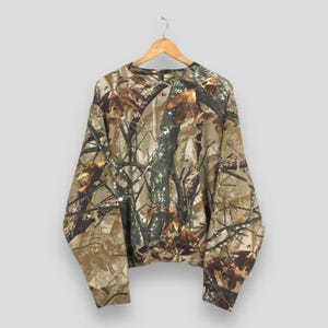 May include: A long-sleeved camouflage sweatshirt hangs from a wooden hanger. The pattern features shades of brown, tan, and green, mimicking a forest environment. The sweatshirt is made of a soft material and has a crew neck.