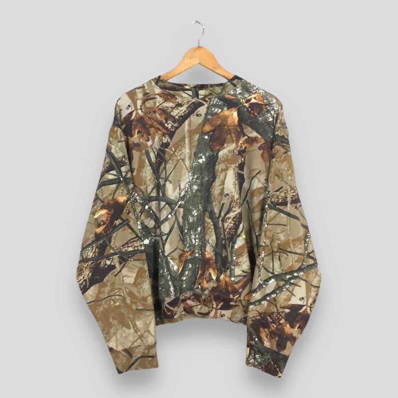 May include: A long-sleeved camouflage sweatshirt hangs from a wooden hanger. The pattern features shades of brown, tan, and green, mimicking a forest environment. The sweatshirt is made of a soft material and has a crew neck.