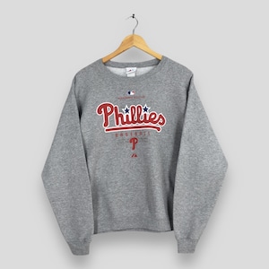 Peut inclure: Un sweat-shirt gris à col rond avec le logo de l'équipe de baseball des Phillies de Philadelphie. Le logo comprend le mot "Phillies" en script rouge avec un contour blanc, deux étoiles bleues et la lettre "P". Le sweat-shirt est suspendu à un cintre en bois.