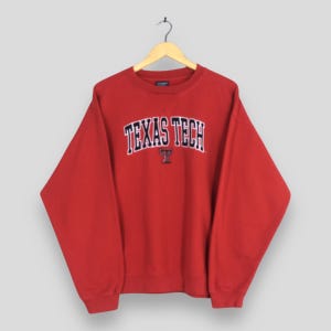 Può includere: Felpa rossa Texas Tech con la scritta "TEXAS TECH" in lettere bianche arcuate sul petto. La felpa ha un girocollo e maniche lunghe. Un piccolo logo "T" è sotto il testo.