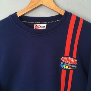 May include: Navy blue sweatshirt with red stripes down the right shoulder and a "DuPont Motorsports" patch. The sweatshirt also features a "Hase Authentic" label on the collar.