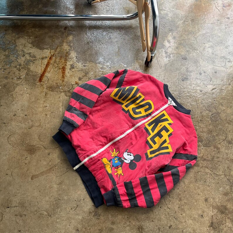 May include: Red zip-up jacket with black and gray striped sleeves. The jacket features the words "MICKEY" in yellow and a graphic of Mickey Mouse. The jacket has a white zipper and black trim.