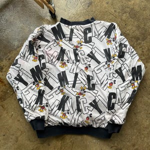 May include: White and black Mickey Mouse sweatshirt. The sweatshirt features a repeating pattern of the word "MICKEY" in large black letters, interspersed with images of Mickey Mouse. The cuffs and neckline are black.