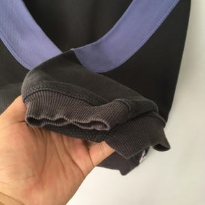 May include: Close-up of a black and gray sweatshirt with a light purple accent across the shoulders. The ribbed cuffs are a darker gray. The fabric has a textured appearance.