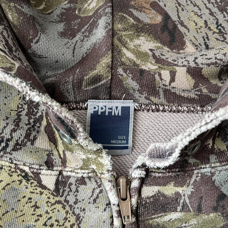 K&ouml;nnte beinhalten: Ein Kapuzen-Sweatshirt mit Camouflage-Muster in Gr&uuml;n-, Braun- und Graut&ouml;nen. Das Etikett lautet "PPFM" mit "SIZE MEDIUM" darunter. Der Rei&szlig;verschluss ist goldfarben.