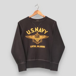 May include: A dark brown U.S. Navy sweatshirt with a yellow emblem featuring an anchor, wings, and the text "U.S. NAVY" and "CLINTON, OKLAHOMA." The crew neck sweatshirt is hanging on a wooden hanger.