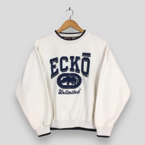 May include: Cream-coloured sweatshirt featuring the brand name "ECKO" in navy blue, outlined in grey, and the word "Unlimited" below a rhino logo. The sweatshirt has navy blue trim around the collar and cuffs.