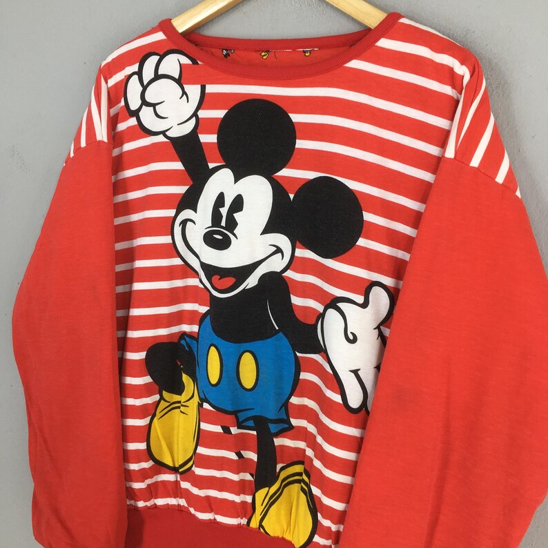 May include: Red and white striped sweatshirt featuring a large graphic of Mickey Mouse. The sleeves are solid red. Mickey Mouse is depicted in his classic pose, wearing blue shorts and yellow shoes. The sweatshirt is hanging on a wooden hanger.