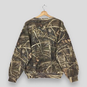 May include: A long-sleeved camouflage sweatshirt with a crew neck, hanging on a wooden hanger. The pattern features shades of brown, green, and tan, resembling a natural outdoor environment. The sweatshirt is made of a soft material.