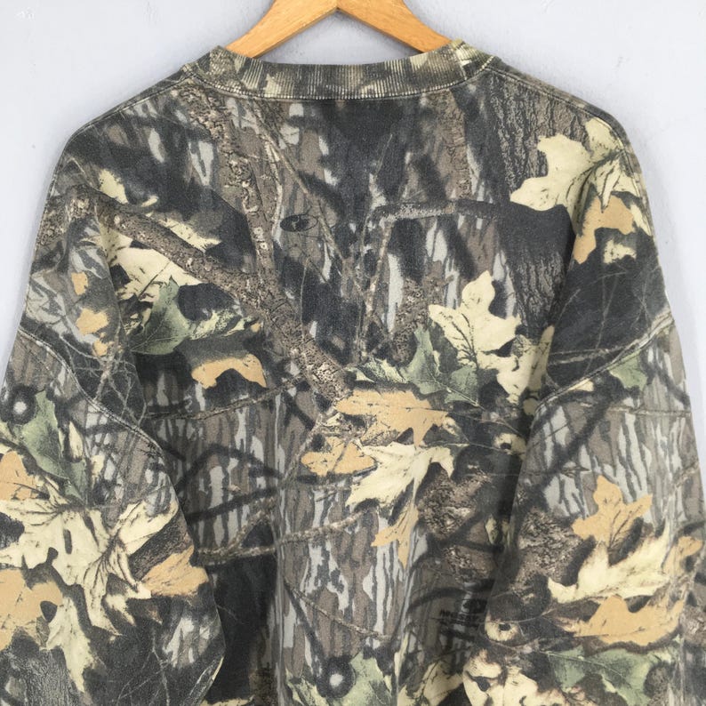 May include: A camouflage-patterned sweatshirt with a crew neck. The design features a mix of brown, green, and tan hues, mimicking a forest environment. The fabric appears to be a medium-weight material, suitable for cooler weather.