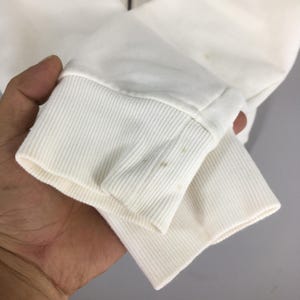 Puede incluir: Primer plano de una manga de sudadera blanca con pu&ntilde;o acanalado. La manga muestra algo de decoloraci&oacute;n. La tela parece ser un material suave y tejido. La imagen est&aacute; tomada sobre un fondo neutro.