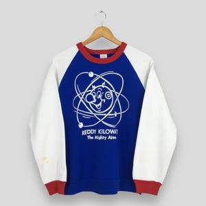 May include: A vintage raglan-sleeve sweatshirt with a blue body, white sleeves, and red trim. The front features a white graphic of an atom with a smiling face and the text "Reddy Kilowatt The Mighty Atom."