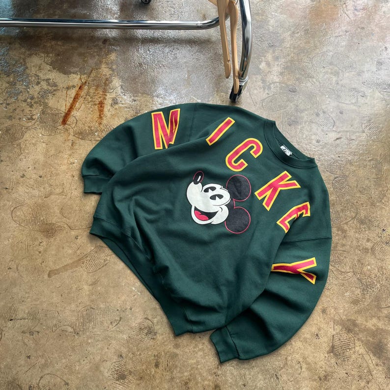 K&ouml;nnte beinhalten: Ein dunkelgr&uuml;nes Sweatshirt mit dem Wort "MICKEY" in roten und gelben Buchstaben. Das Sweatshirt hat eine Mickey Mouse-Grafik. Das Kleidungsst&uuml;ck ist aus einem weichen Material gefertigt.