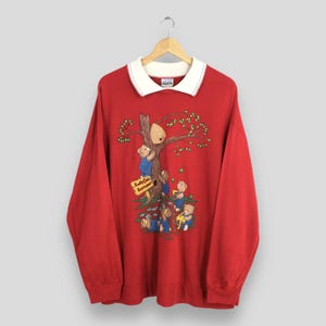 Może przedstawiać: Czerwona bluza z długim rękawem i białym kołnierzem. Z przodu znajduje się ilustracja misiów wspinających się na drzewo z ulem. Znak głosi "Honey Cake Bottom".