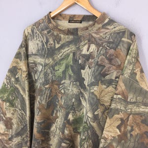 K&ouml;nnte beinhalten: Ein lang&auml;rmeliges Camouflage-Shirt mit Rundhalsausschnitt. Das Shirt zeigt ein Muster aus braunen, gr&uuml;nen und beigen Bl&auml;ttern und &Auml;sten. Das Wort "COMMANDER" ist auf dem Kragen aufgedruckt. Das Shirt h&auml;ngt an einem Holzb&uuml;gel.