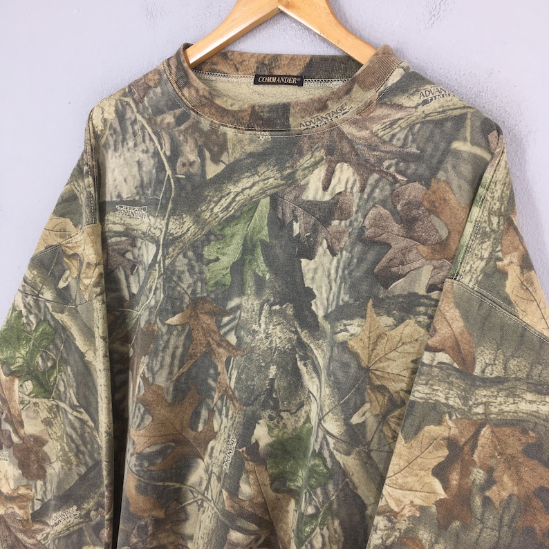 K&ouml;nnte beinhalten: Ein lang&auml;rmeliges Camouflage-Shirt mit Rundhalsausschnitt. Das Shirt zeigt ein Muster aus braunen, gr&uuml;nen und beigen Bl&auml;ttern und &Auml;sten. Das Wort "COMMANDER" ist auf dem Kragen aufgedruckt. Das Shirt h&auml;ngt an einem Holzb&uuml;gel.