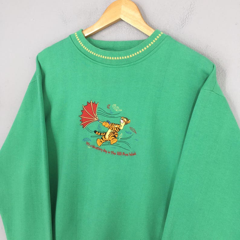 May include: Green sweatshirt featuring an embroidered image of Tigger from Winnie the Pooh holding an umbrella in the wind. The text "It's a Whiffletry day in The 100 Aker Wood" is below the image. The sweatshirt has a yellow and white striped collar.