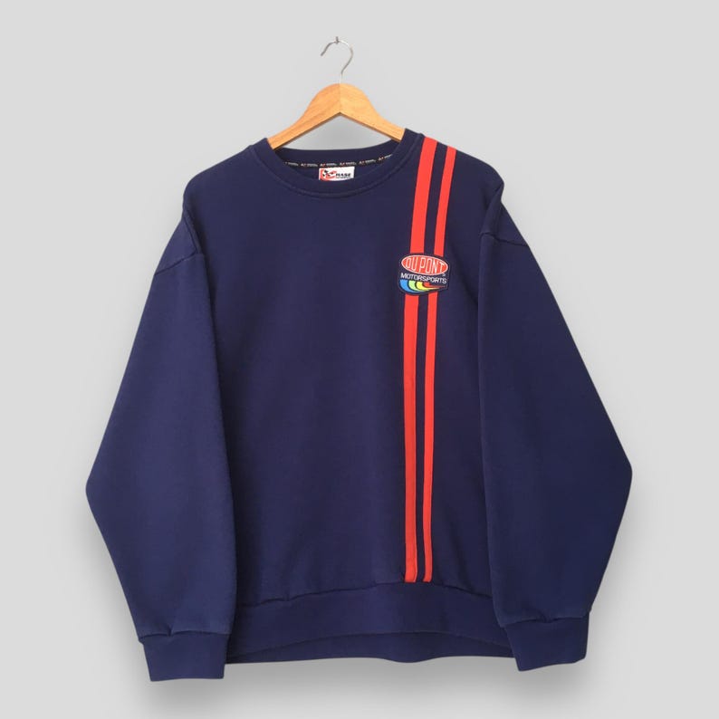 May include: Navy blue sweatshirt with red vertical stripes on the left side. The sweatshirt features the "DuPont Motorsports" logo in the upper right corner. The sweatshirt is hanging on a wooden hanger.