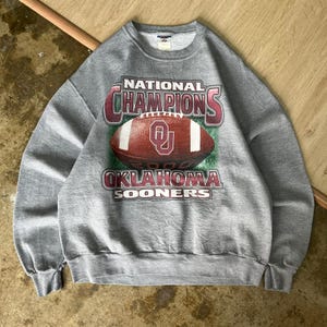 May include: A gray sweatshirt featuring a football graphic with the text "NATIONAL CHAMPIONS 2000 OKLAHOMA SOONERS." The graphic includes a football with the OU logo. The sweatshirt has a crew neck and ribbed cuffs.