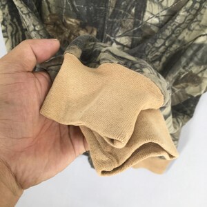May include: A camouflage-patterned jacket with a tan-colored ribbed cuff. The jacket features a zipper and a woodland camouflage design in shades of green, brown, and gray. The cuff is made of a soft, knitted material.