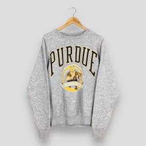 May include: A grey Purdue University sweatshirt. The word "PURDUE" is arched across the chest in black with yellow accents. Below the word "PURDUE" is a circular graphic with a cartoon figure and the words "PURDUE UNIVERSITY" and "BOILERMAKERS".