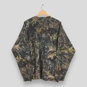 Puede incluir: Una sudadera de manga larga con estampado de camuflaje y cuello redondo. El dise&ntilde;o presenta una mezcla de marr&oacute;n, verde y negro, que recuerda a una escena forestal. La sudadera est&aacute; colgada de una percha de madera.