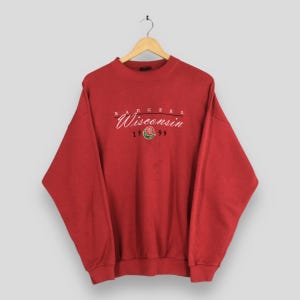 May include: A red sweatshirt with the text "BADGERS Wisconsin 1999" embroidered in white and green. The sweatshirt is hanging on a wooden hanger against a light background. The design includes a rose graphic.