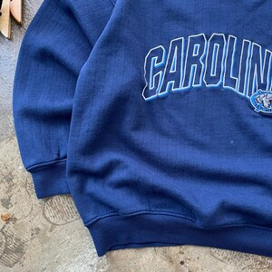 May include: A navy blue long-sleeve sweatshirt featuring a vertical ribbed texture. The front displays the word CAROLINA in large white block letters with a light blue outline, accompanied by a small circular graphic featuring a ram mascot logo.