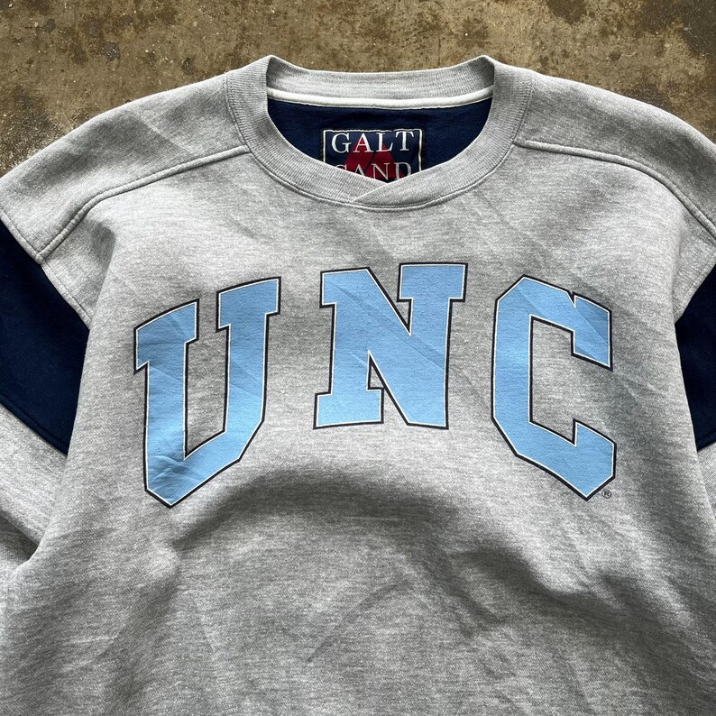 Puede incluir: Una sudadera gris con las letras "UNC" en azul claro en la parte delantera. La sudadera tiene detalles en azul marino en las mangas y un peque&ntilde;o logotipo "GALT AND" cerca del cuello. La prenda est&aacute; hecha de un tejido suave.