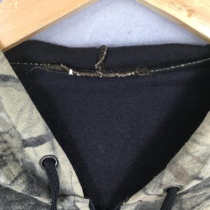 May include: Close-up of a camouflage-patterned hoodie. The hood is lined with a dark navy blue fabric. The drawstring is black, and the eyelets are silver. The hoodie is hanging on a wooden hanger.