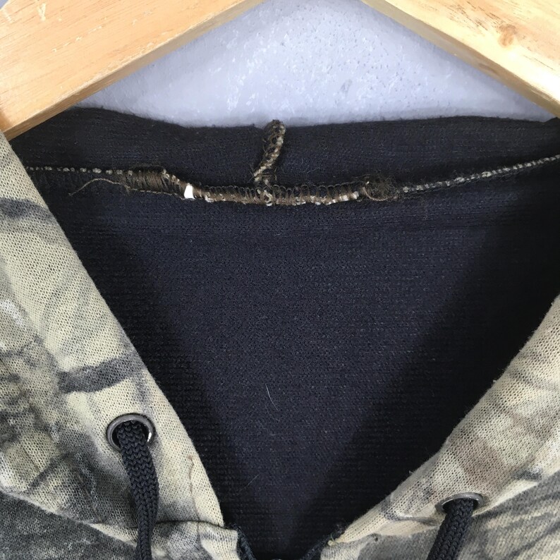 May include: Close-up of a camouflage-patterned hoodie. The hood is lined with a dark navy blue fabric. The drawstring is black, and the eyelets are silver. The hoodie is hanging on a wooden hanger.