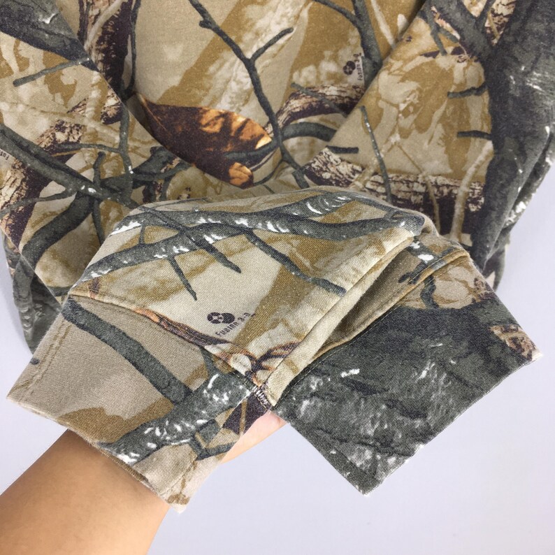 May include: A camouflage-patterned sweatshirt with a hood. The fabric features a blend of brown, tan, and green hues, mimicking a forest environment. The sleeve cuff is visible, showcasing the detailed print and texture of the material.