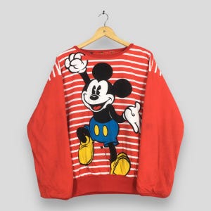 May include: Red and white striped sweatshirt featuring a large graphic of Mickey Mouse. The sleeves are solid red. Mickey is wearing blue shorts and yellow shoes, with his arms raised in a cheerful pose. The sweatshirt is hanging on a wooden hanger.