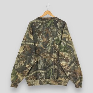 K&ouml;nnte beinhalten: Ein Sweatshirt mit Camouflage-Muster h&auml;ngt an einem Holzb&uuml;gel. Das lang&auml;rmlige Oberteil weist eine Mischung aus Braun-, Gr&uuml;n- und Beiget&ouml;nen auf, die eine Waldumgebung nachahmen. Das Sweatshirt besteht aus einem weichen Material und hat einen Rundhalsausschnitt.