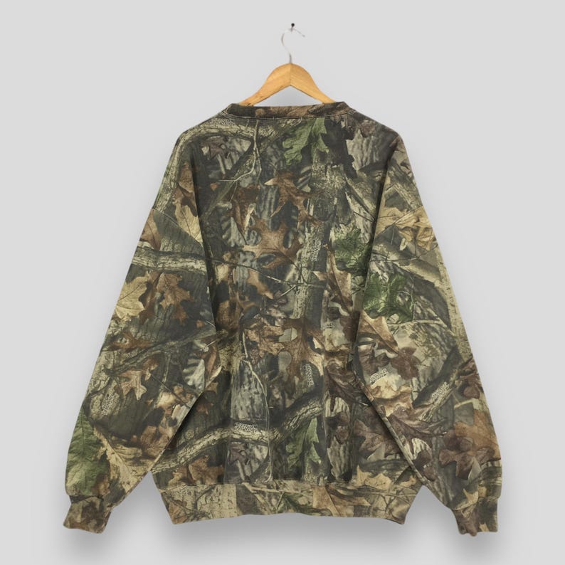 K&ouml;nnte beinhalten: Ein Sweatshirt mit Camouflage-Muster h&auml;ngt an einem Holzb&uuml;gel. Das lang&auml;rmlige Oberteil weist eine Mischung aus Braun-, Gr&uuml;n- und Beiget&ouml;nen auf, die eine Waldumgebung nachahmen. Das Sweatshirt besteht aus einem weichen Material und hat einen Rundhalsausschnitt.