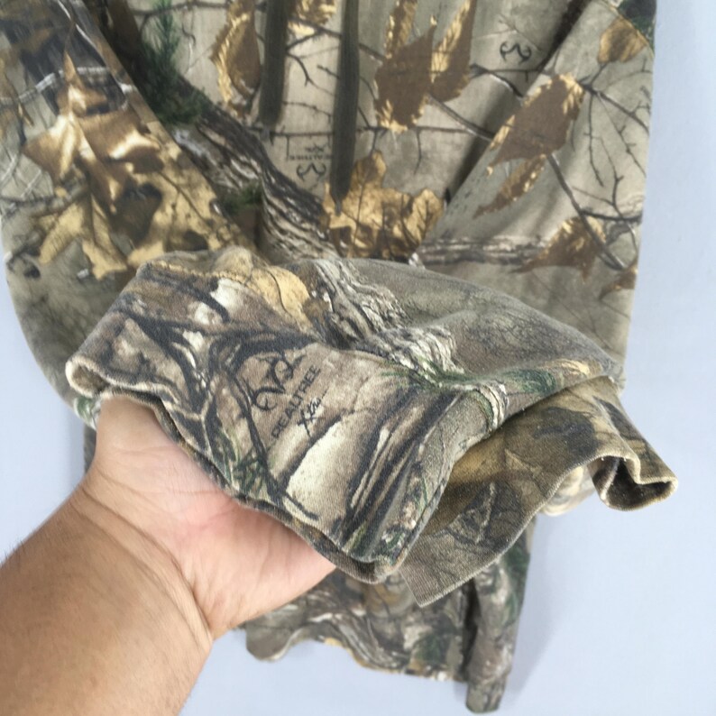 Op de afbeelding: Hoodie met camouflagepatroon en een trekkoord. De stof heeft een mix van bruine, groene en bruine tinten, die een bosrijke omgeving nabootsen. De manchet toont het Realtree-logo. Het ontwerp suggereert outdoorkleding of jachtkleding.