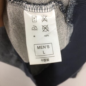 May include: Close-up of a clothing tag with care symbols and text. The tag reads "MEN'S L" and "100%" with Chinese characters. The tag is attached to a blue and gray fabric.