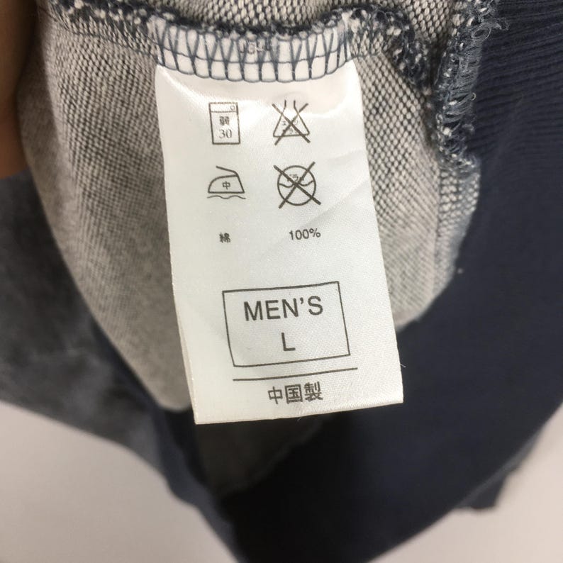 May include: Close-up of a clothing tag with care symbols and text. The tag reads "MEN'S L" and "100%" with Chinese characters. The tag is attached to a blue and gray fabric.