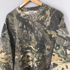 May include: A camouflage-patterned sweatshirt in shades of green, brown, and tan. The crew neck sweatshirt is hanging on a wooden hanger. The fabric features a detailed print of leaves and branches.