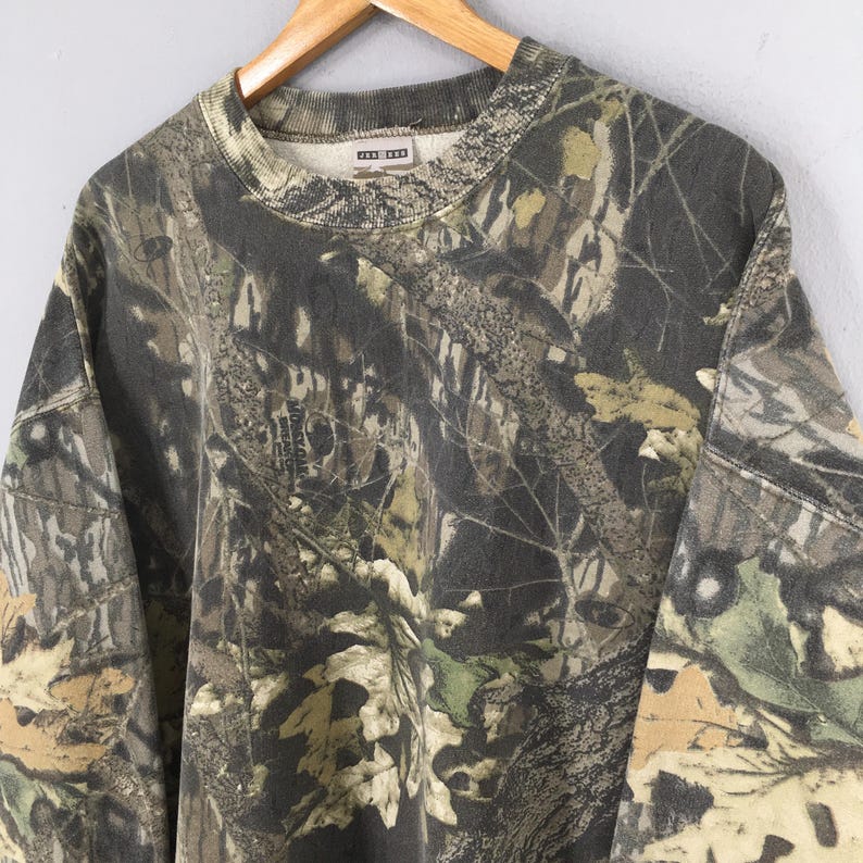 May include: A camouflage-patterned sweatshirt in shades of green, brown, and tan. The crew neck sweatshirt is hanging on a wooden hanger. The fabric features a detailed print of leaves and branches.