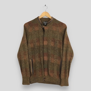 May include: A vintage, button-up cardigan in shades of olive green, brown, and rust. The sweater features a textured, plaid-like pattern, a stand-up collar, and two front pockets with leather trim. The garment is hanging on a wooden hanger.