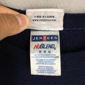 Puede incluir: Primer plano de una etiqueta de sudadera azul marino con las marcas "JERZEES" y "NuBlend" en rojo y blanco. La etiqueta tambi&eacute;n incluye el texto "1-888-RJ-SURFS" y "www.ronjons.com". La etiqueta indica que la prenda est&aacute; hecha en Honduras.
